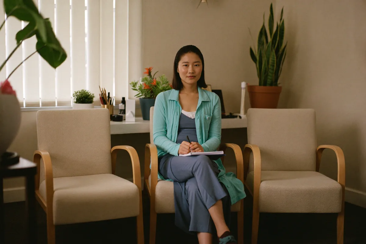 A counsellor seated in a calm consulting room with two armchairs and soft light, notebook on her lap