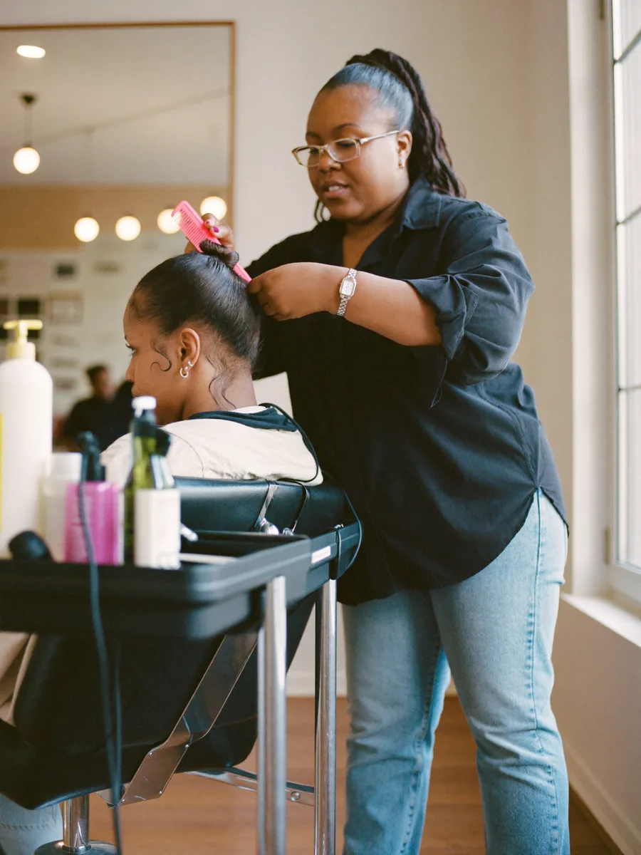 A Black hair stylist sectioning a client's hair in a salon chair, colour trolley beside her