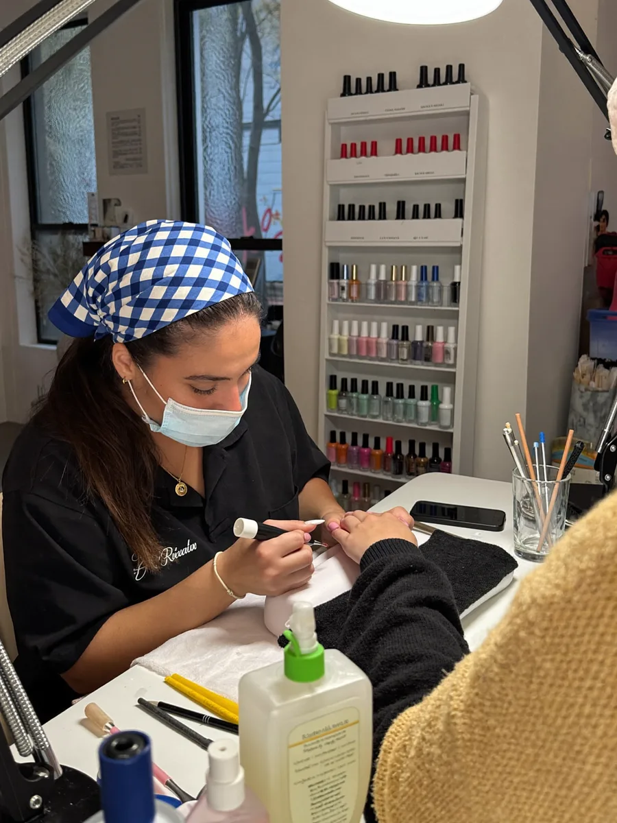 A Latina nail artist painting a client's nails at a manicure table, gel lamps and polish rack in view