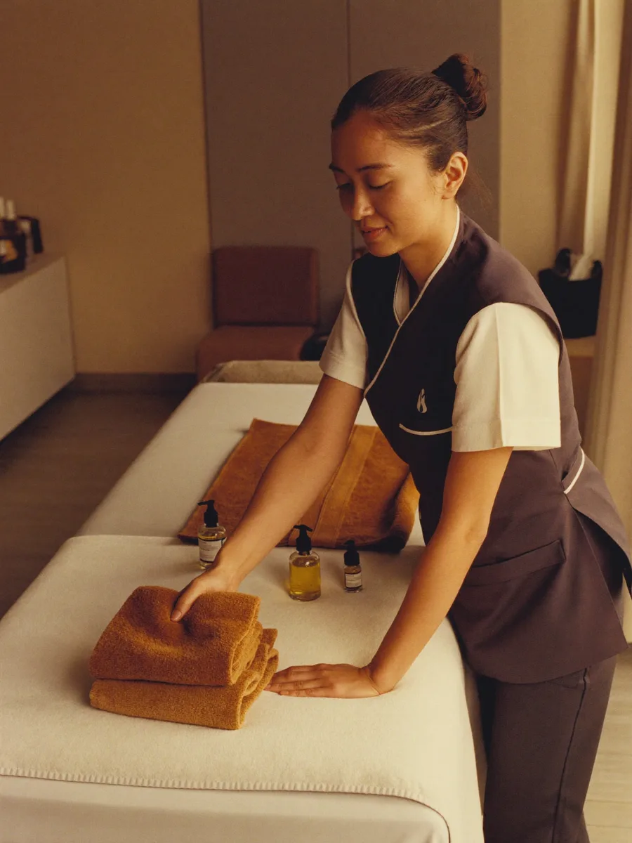 A spa therapist preparing a treatment room, folded towels and oils laid out beside the massage table