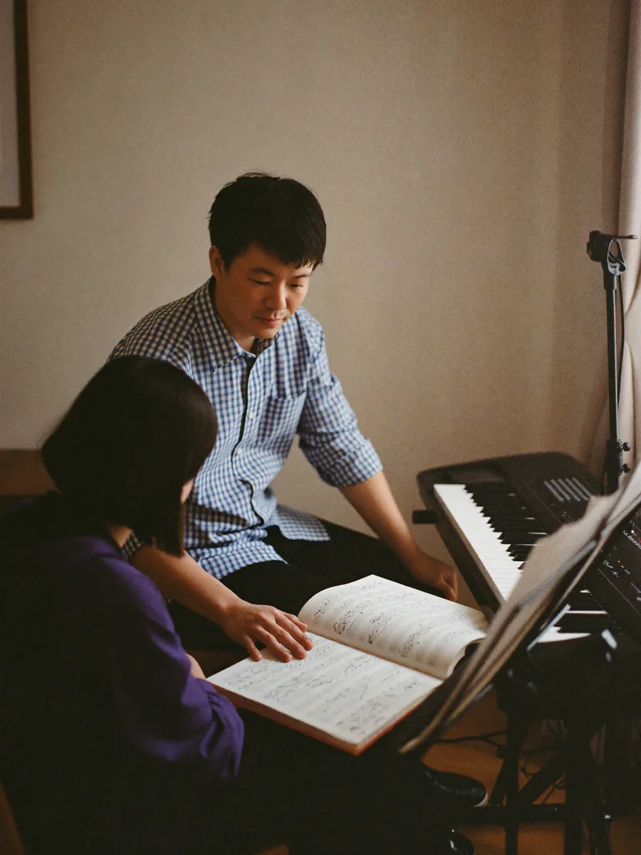 An East Asian music teacher beside a student at a keyboard in a home studio, notation book open