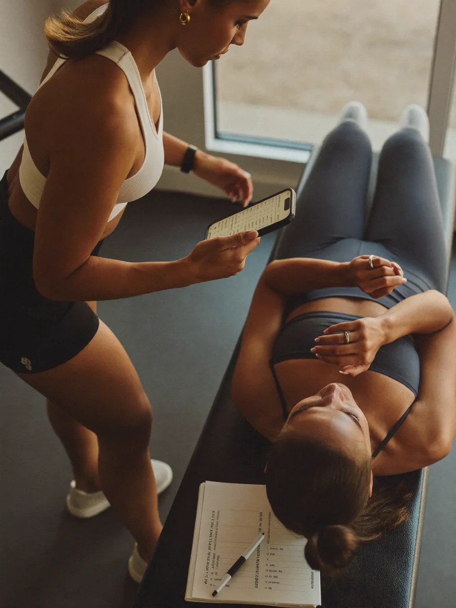 A personal trainer checking the session plan on her phone while a client rests on the bench between sets