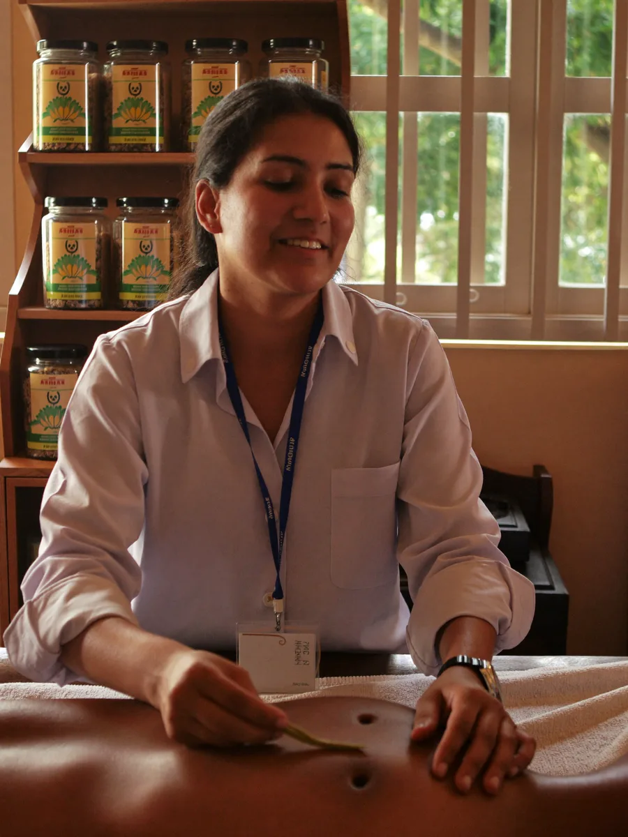 A wellness practitioner giving a herbal oil treatment, jars of preparations on the shelf behind her